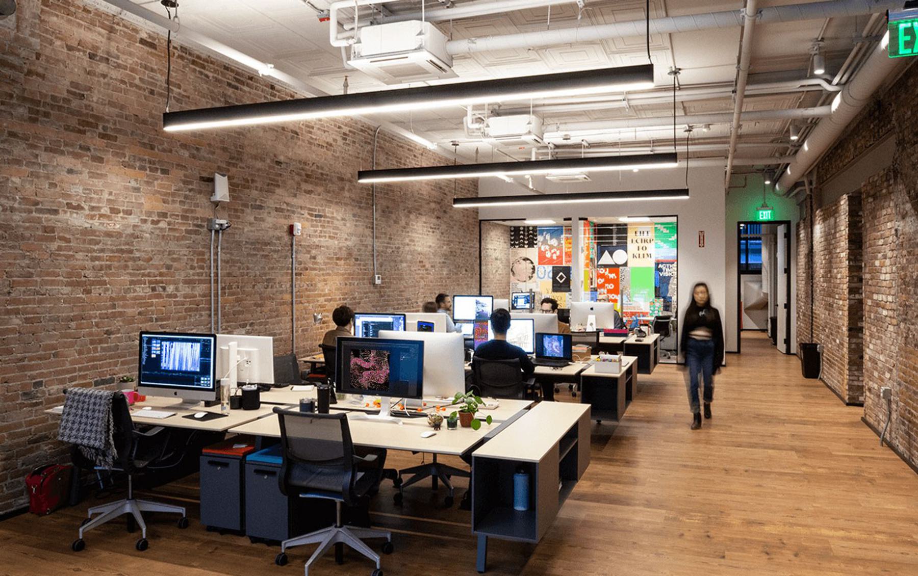 A modern office space with exposed brick walls features several people working at their desk and an individual walking down the central aisle.