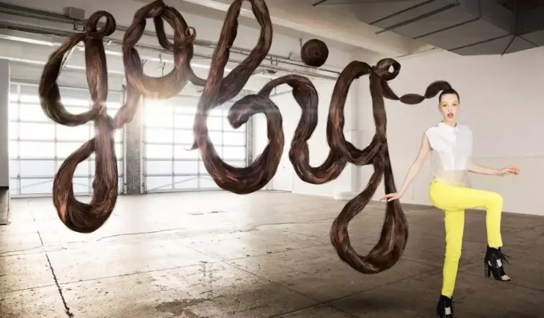 A woman in bright yellow pants stands energetically in a large industrial space. Her long brown hair is styled to spell out "GO BIG" in mid-air.