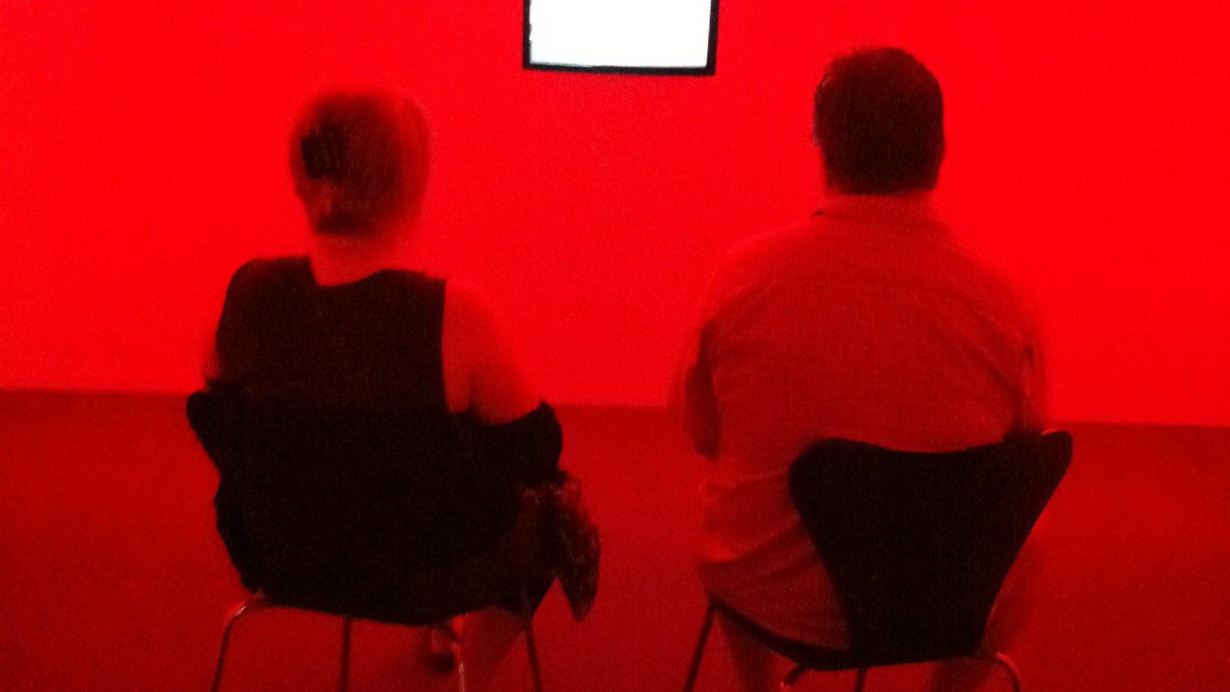 Two people sitting on chairs in front of a white screen in a small room bathed in red light.