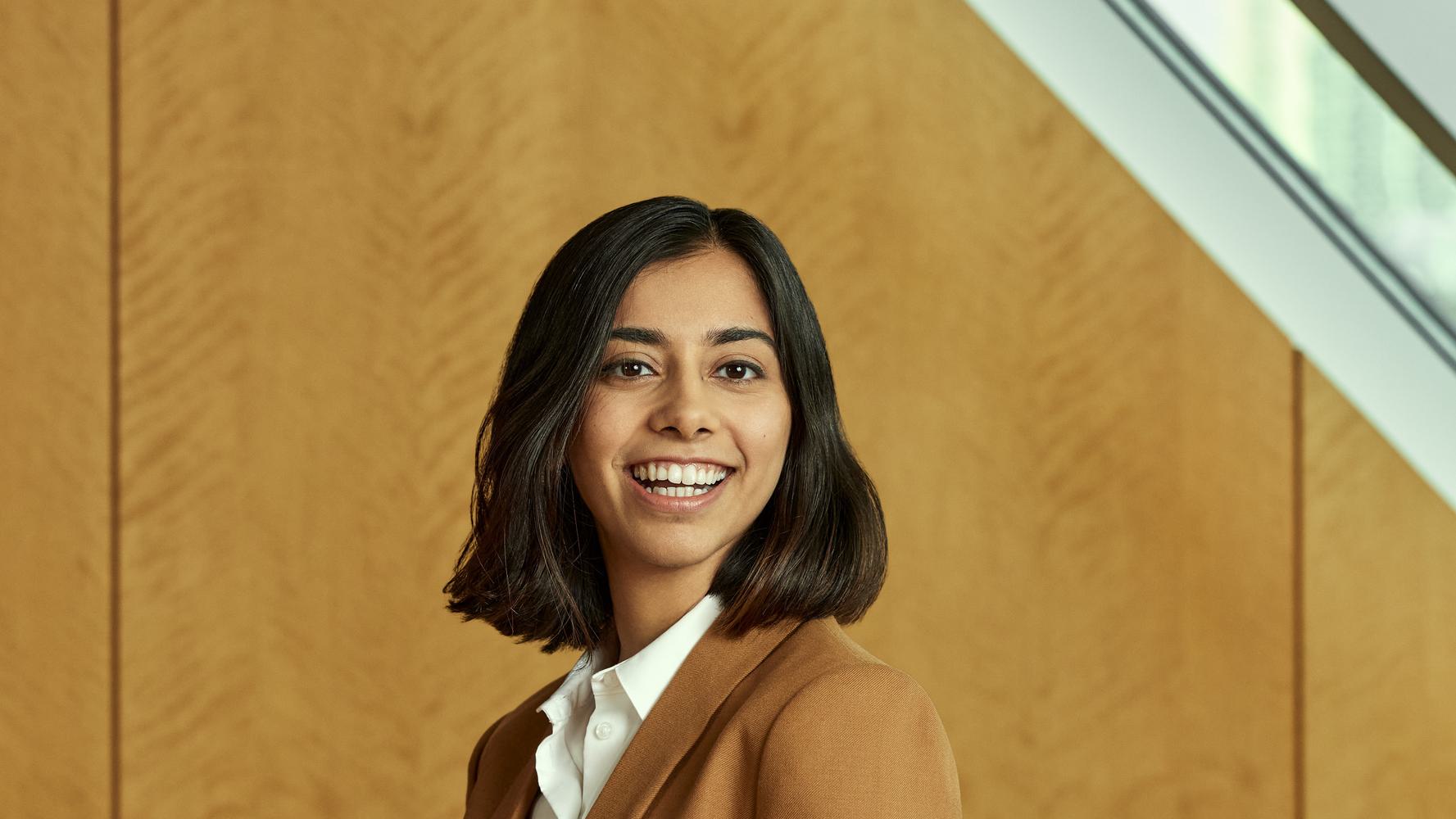 A smiling person with shoulder-length dark hair is wearing a white shirt and a tan blazer. They are standing in front of a wooden wall with a slanted window above.