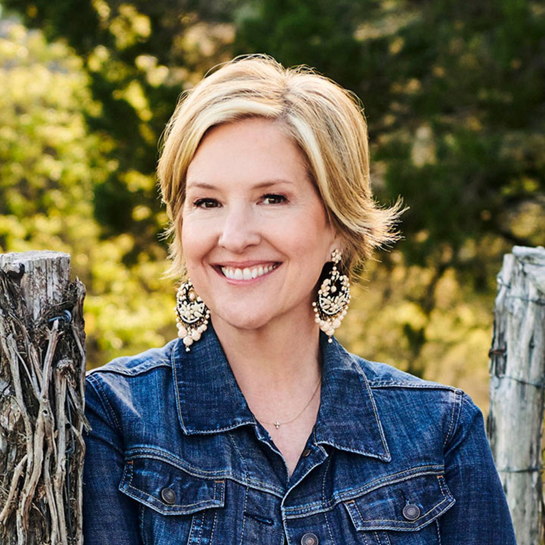 A woman with short blonde hair smiles warmly at the camera. She is wearing a blue denim jacket and large, ornate earrings. She stands next to an aged wooden fence, with a blurred background of green foliage and dappled sunlight.