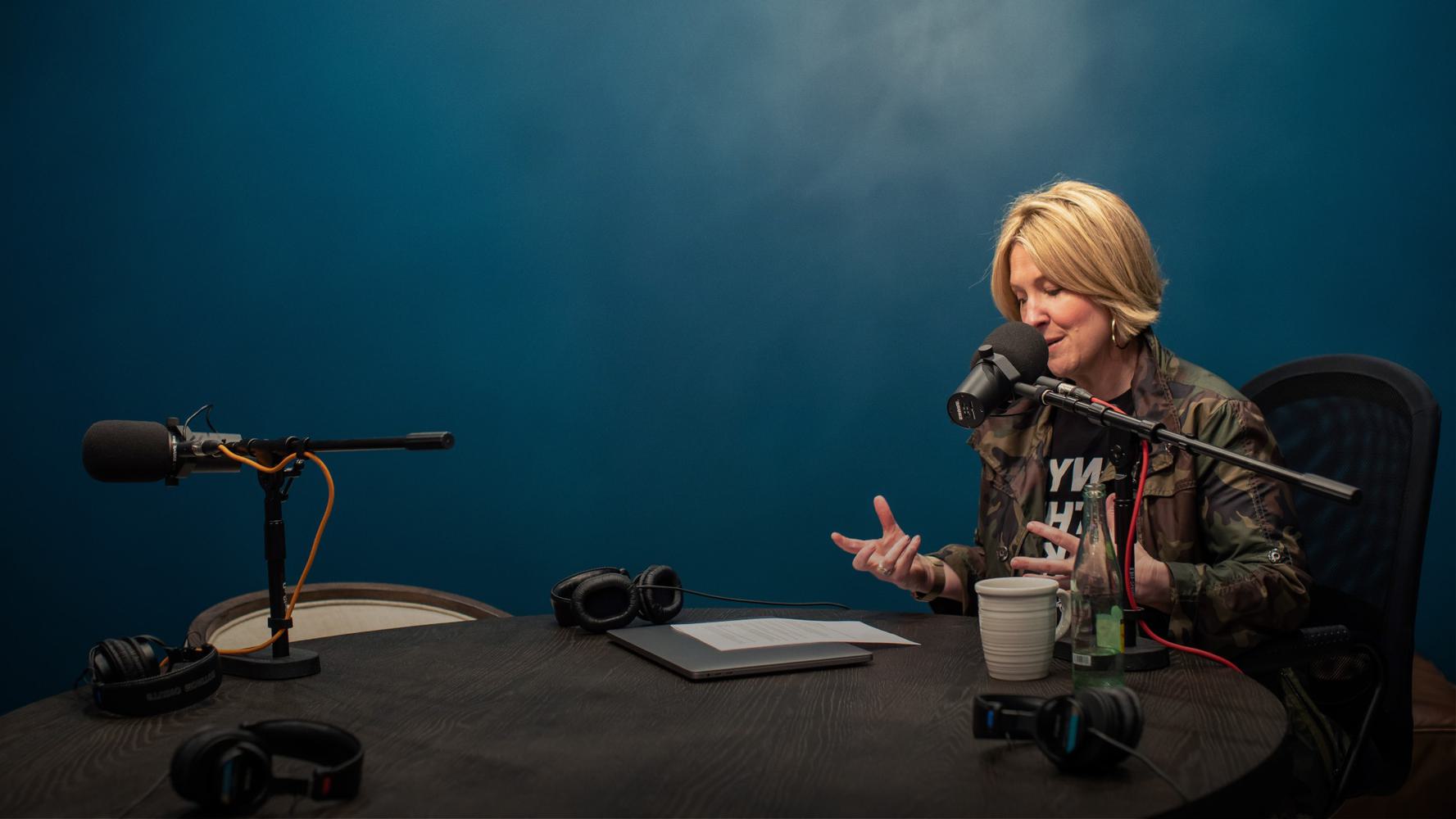 A woman sits at a round table in a podcast studio. She is speaking into a microphone on a boom arm and gesturing with her hands.