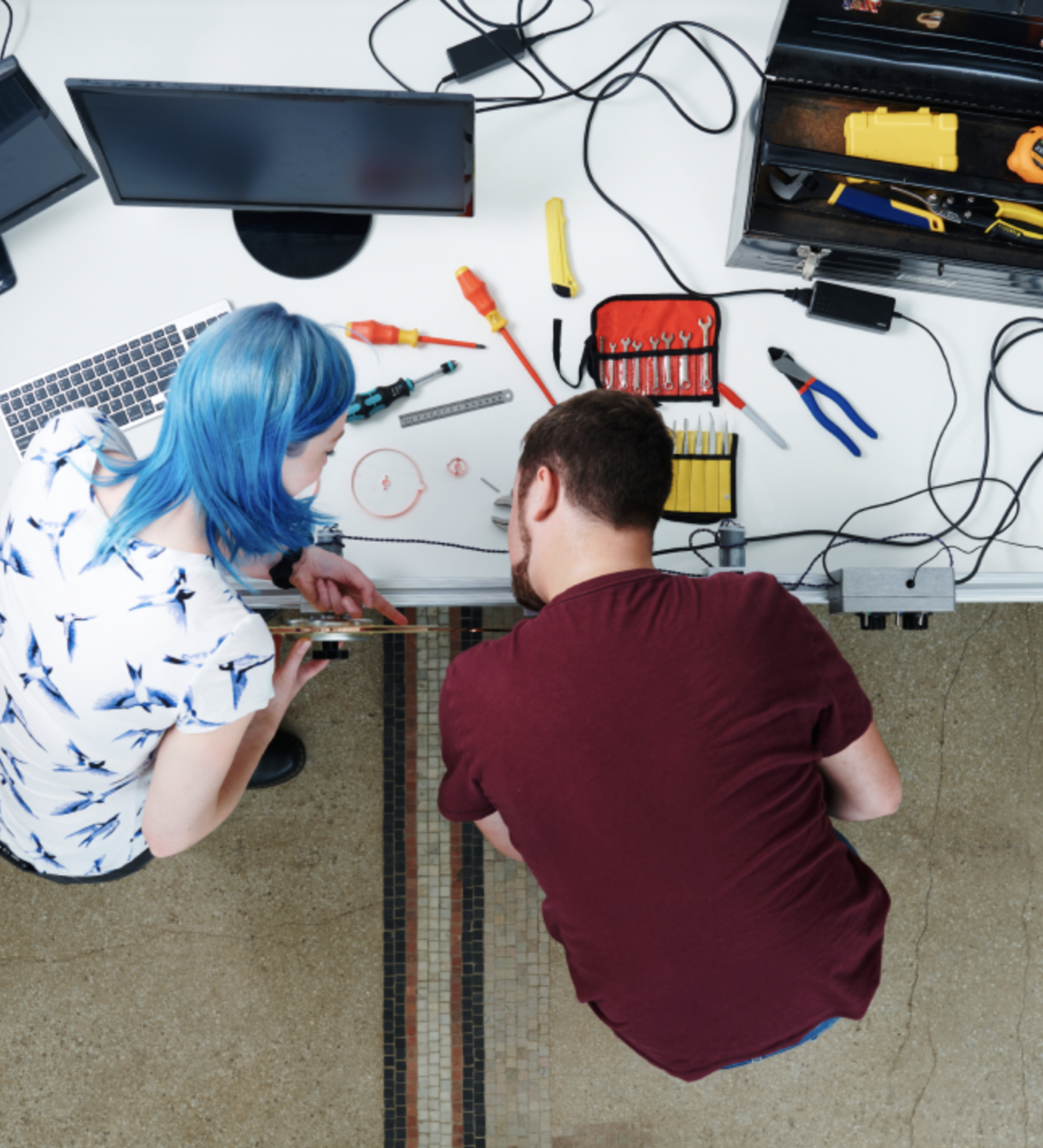 Two technicians work together at a desk with various tools and electronics.