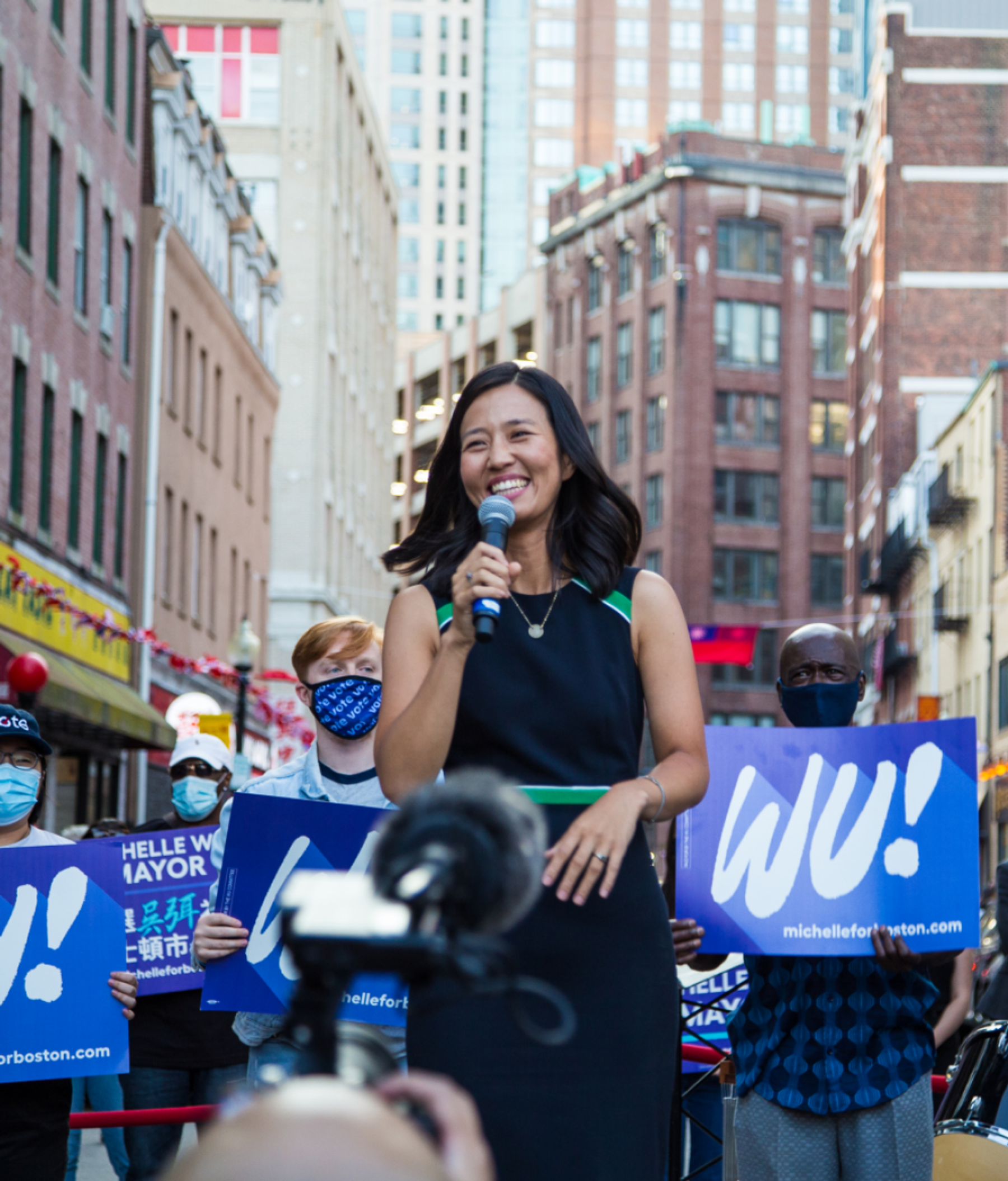 Michelle Wu speaks into a microphone at an outdoor event in front of a crowd, some of whom hold blue "Wu!" signs. The event is set in a street in Boston with tall buildings in the background.