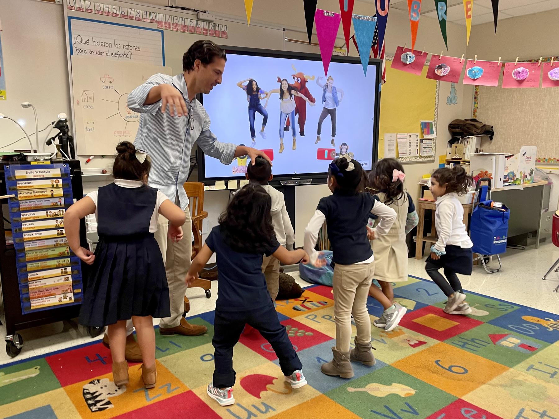 An adult and elementary school students are engaged in a dance activity in a colorful classroom. They mimic the movements of people on a screen at the front of the room.