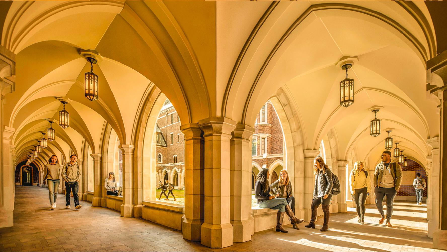 Students walking, talking, and sitting under a series of brick archways on a sunny day.