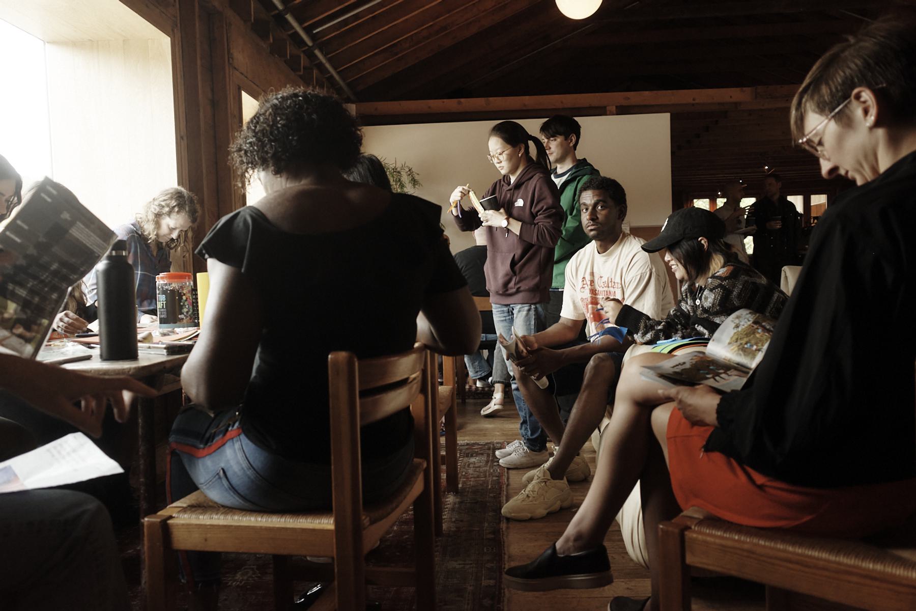A group of people are sitting and standing in a wooden room with a high ceiling engaged in crafts and reading magazines.